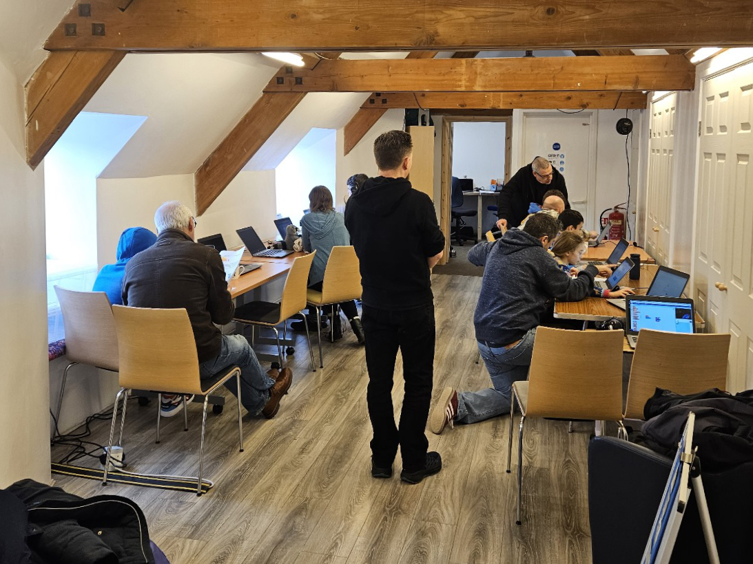 A picture of our Code Club in full flow at Jigsaw Thornbury! There are two banks of desks lining the outside of the room, six children are sat at laptops being guided by volunteers and parents.